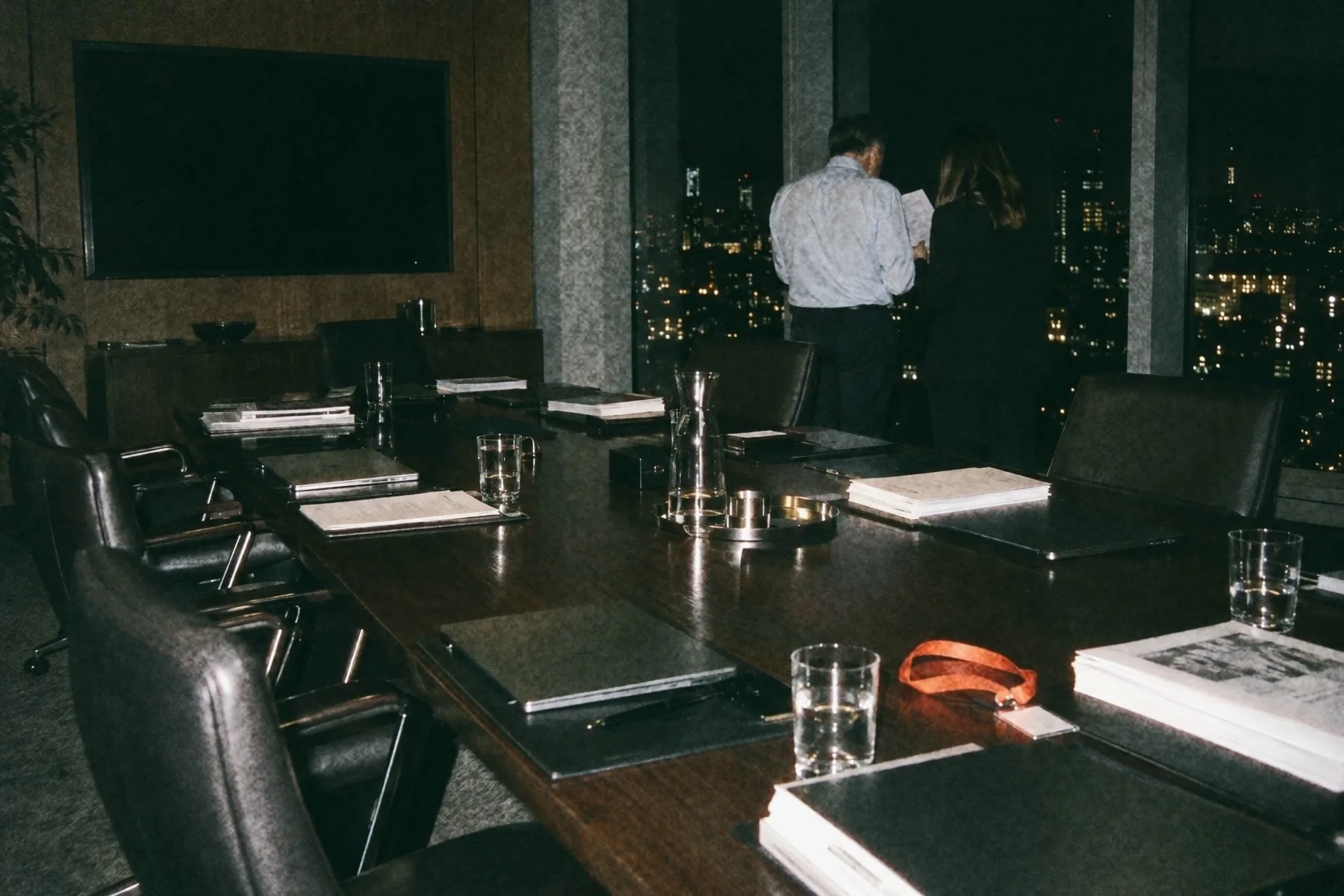 A boardroom on a high floor late in the evening, chairs pushed back after a meeting, two people by the window going over one page together and an orange lanyard on the table