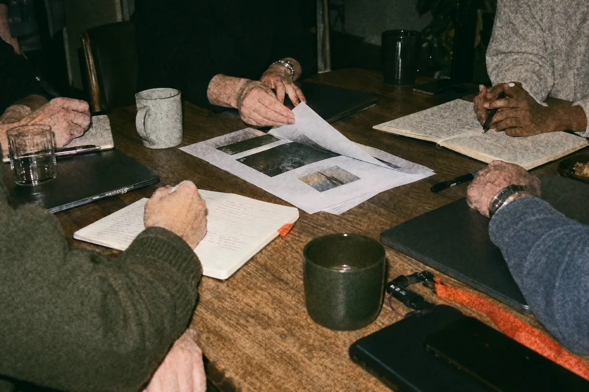 Hands around a table in the evening, one person turning a printed page so the others can read it