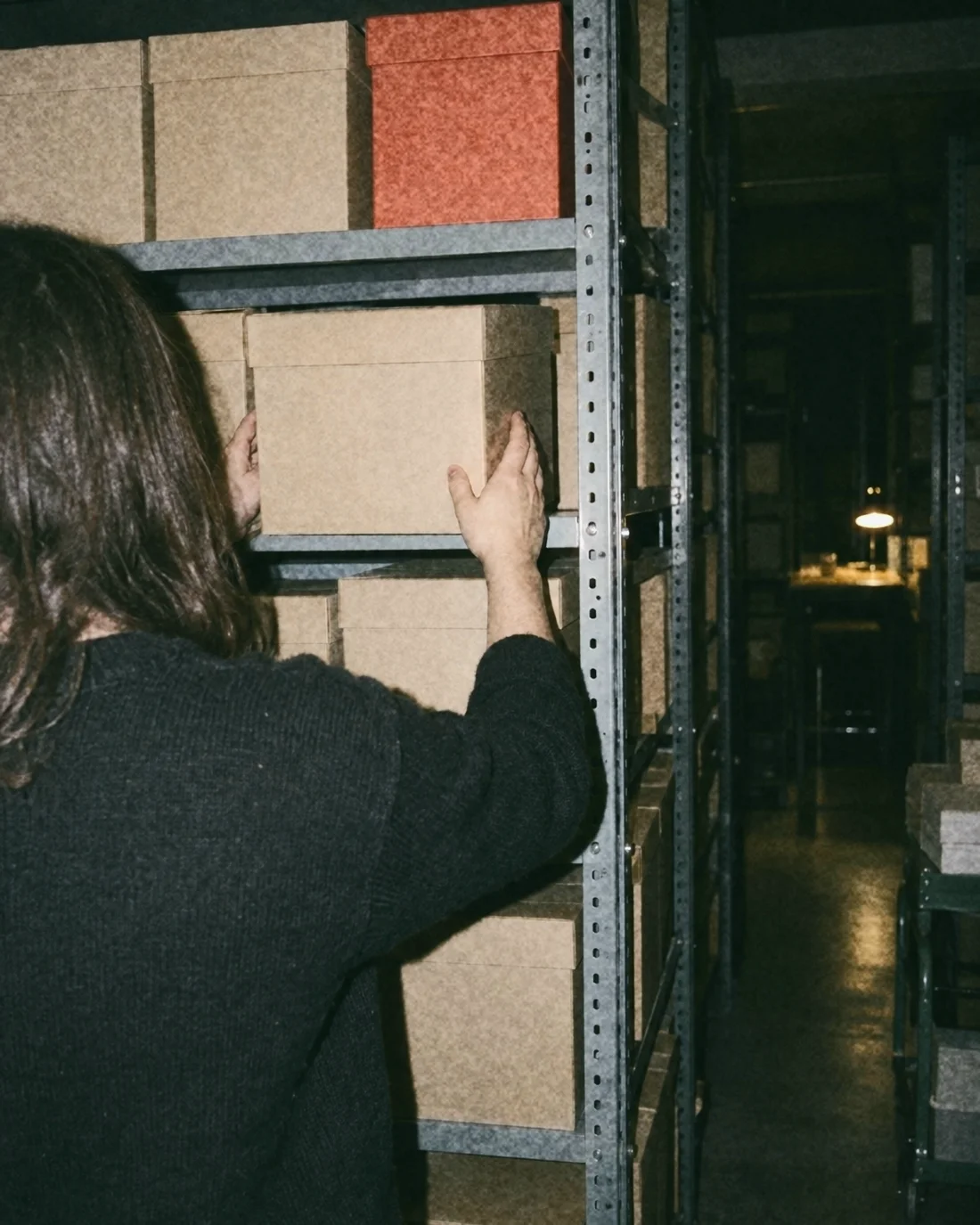 A hand returning a box to an archive shelf of plain boxes, one orange box among them
