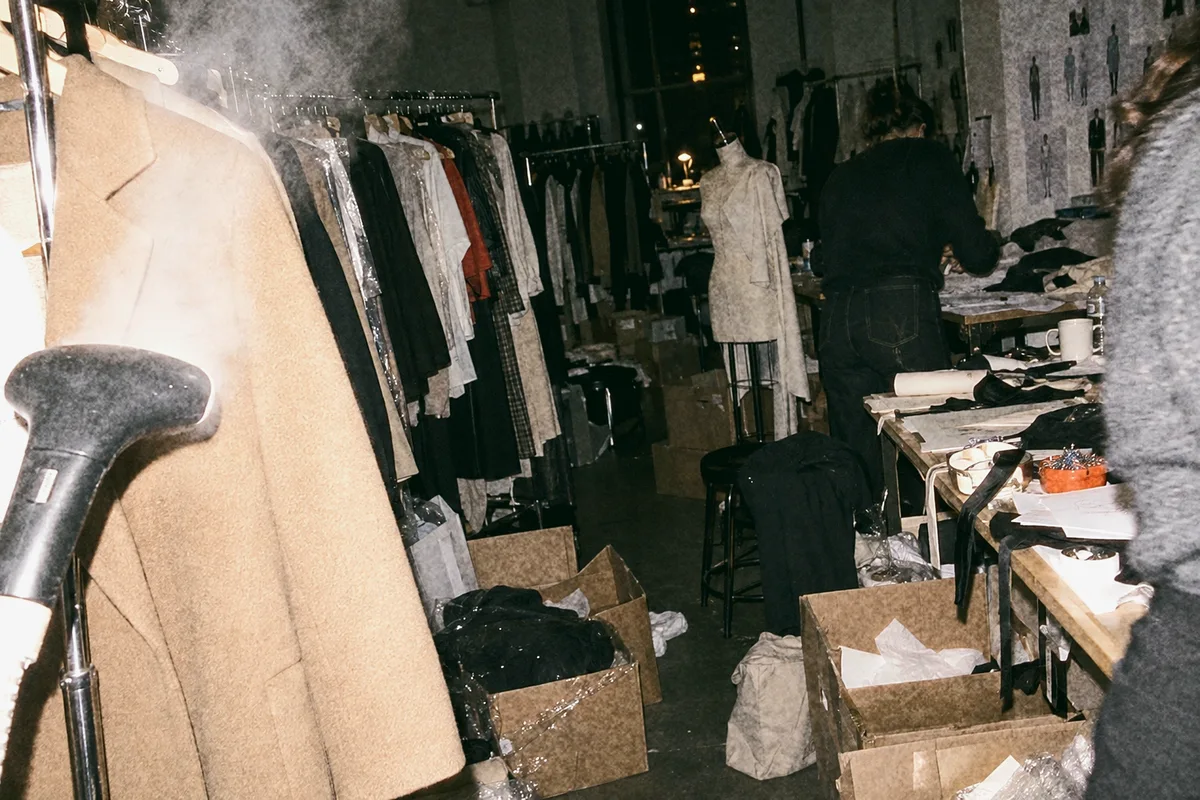 Steam from a garment steamer lit by flash in a crowded studio, a coat on a hanger and a rail of samples behind