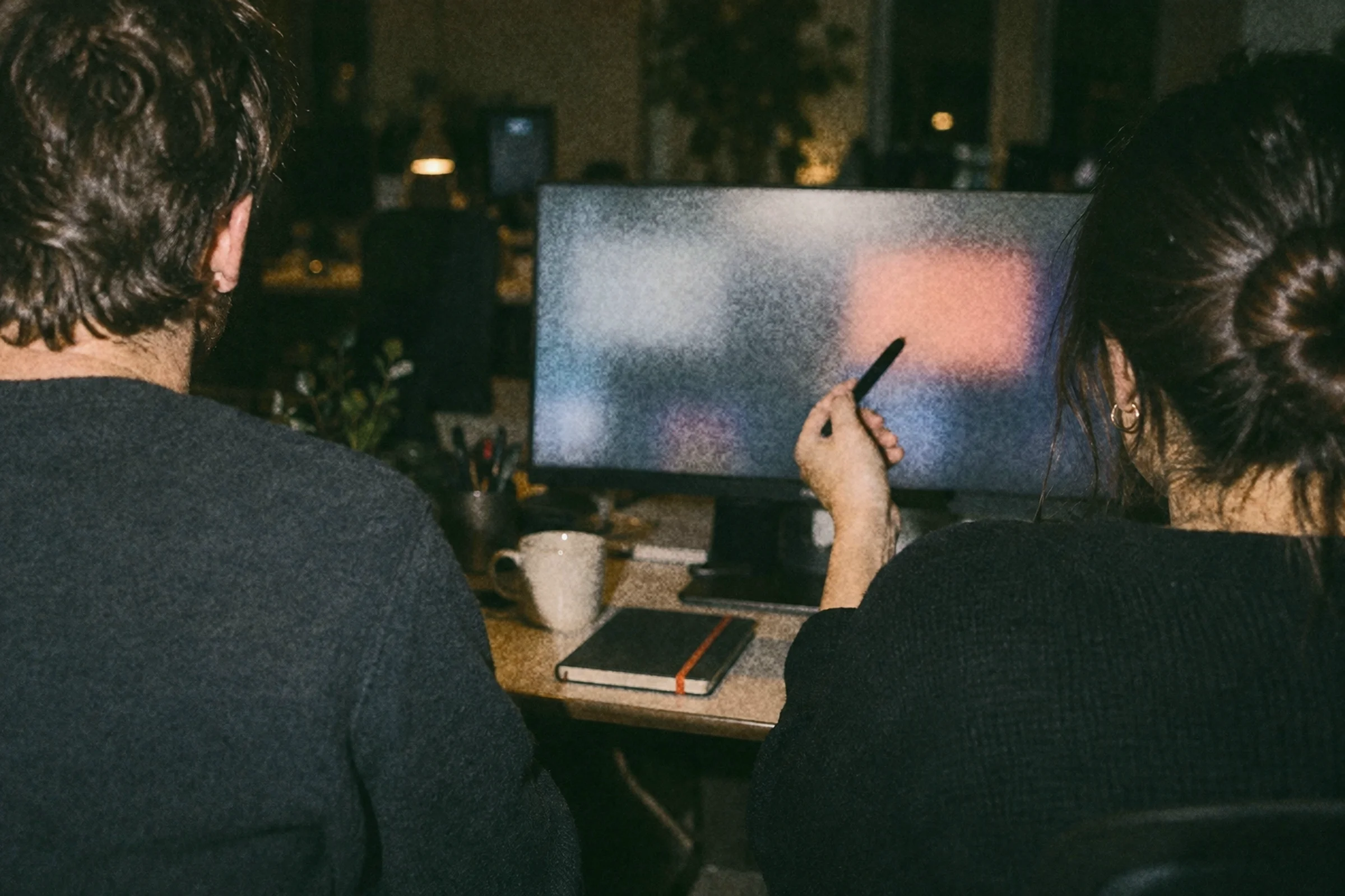 Two people at a desk in the evening, one pointing at a screen while the other takes notes